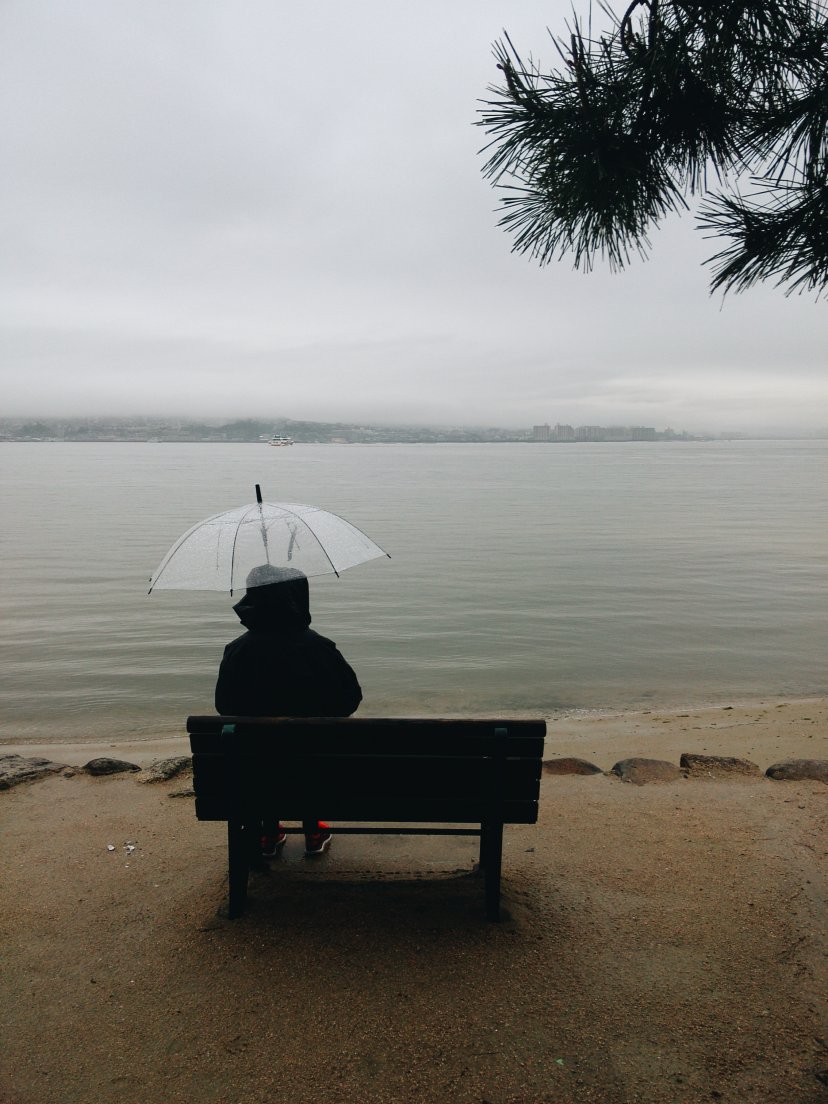 A woman sat on a bench in front of the sea with a transparent umbrella on a rainy day