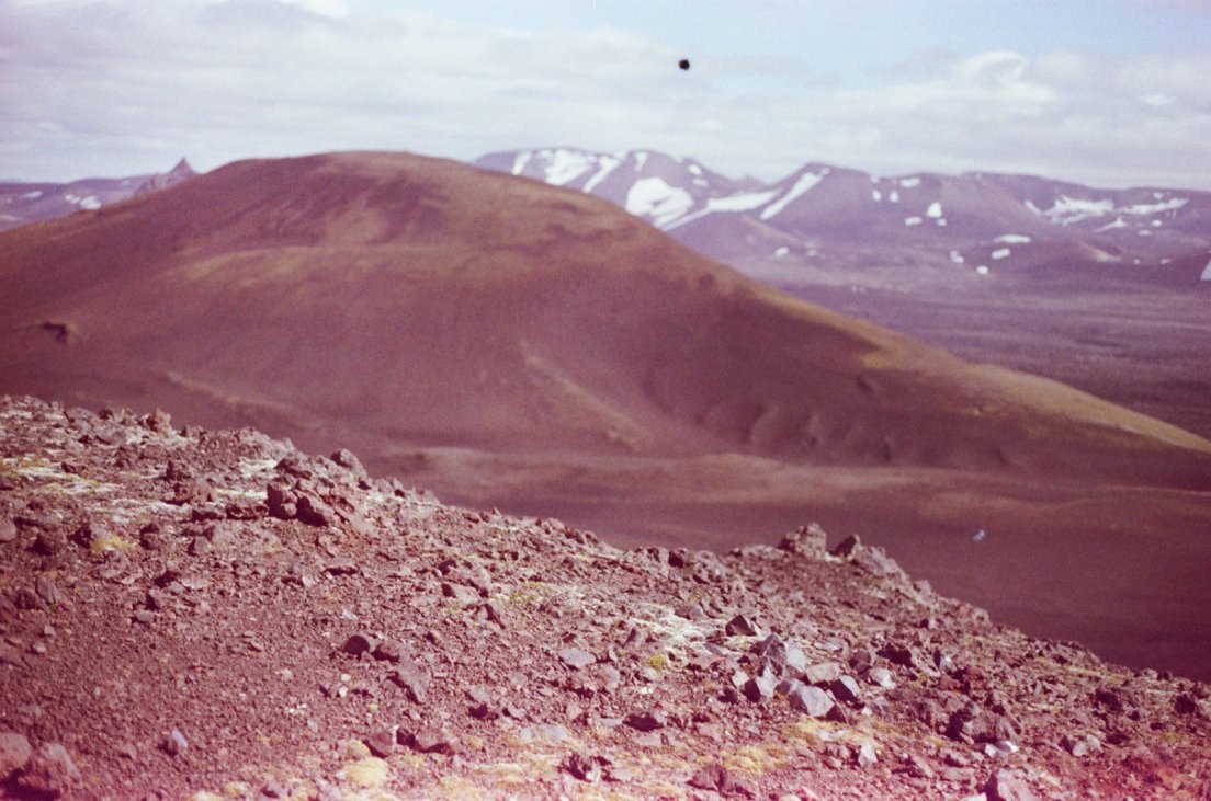 Magenta-tinted photograph of the surroundings of the volcano with lava rocks and snowy-capped mountains