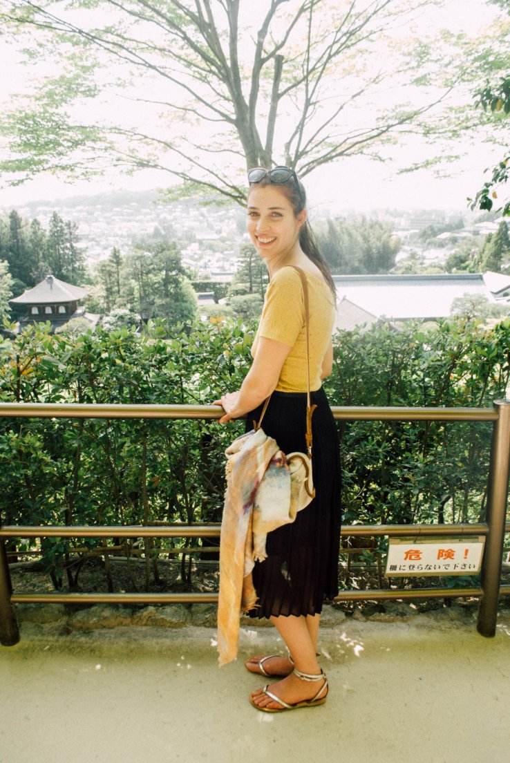 Western girl with mustard top and black skirt smiling in front of the rooftops of the temple as seen from uphill