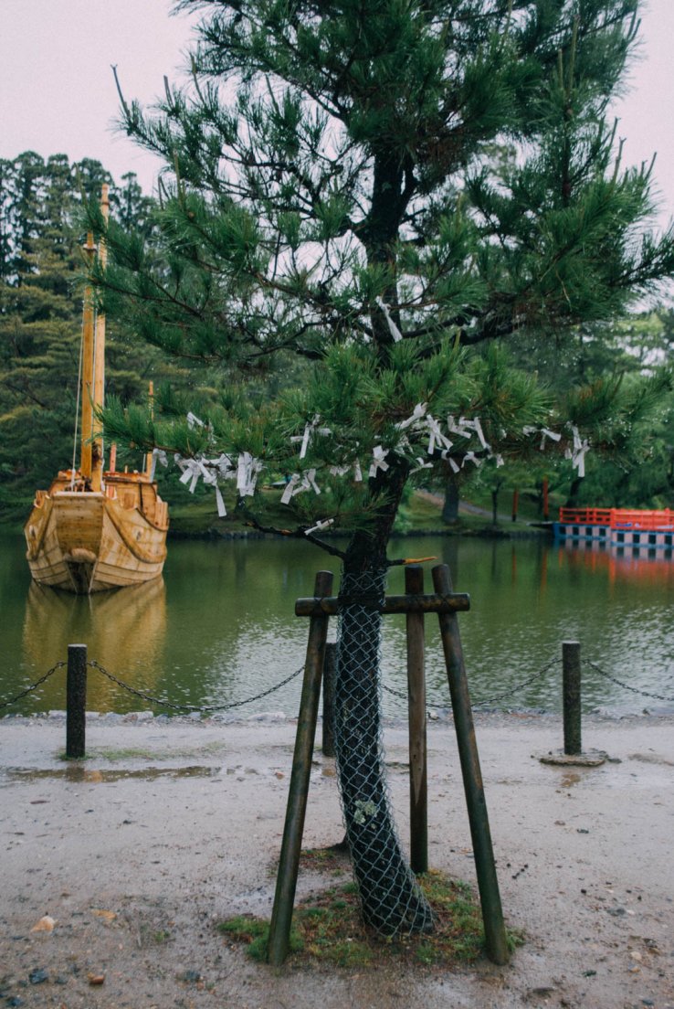 A tree covered in paper wishes in front of a small lake with a pretty barge floating there