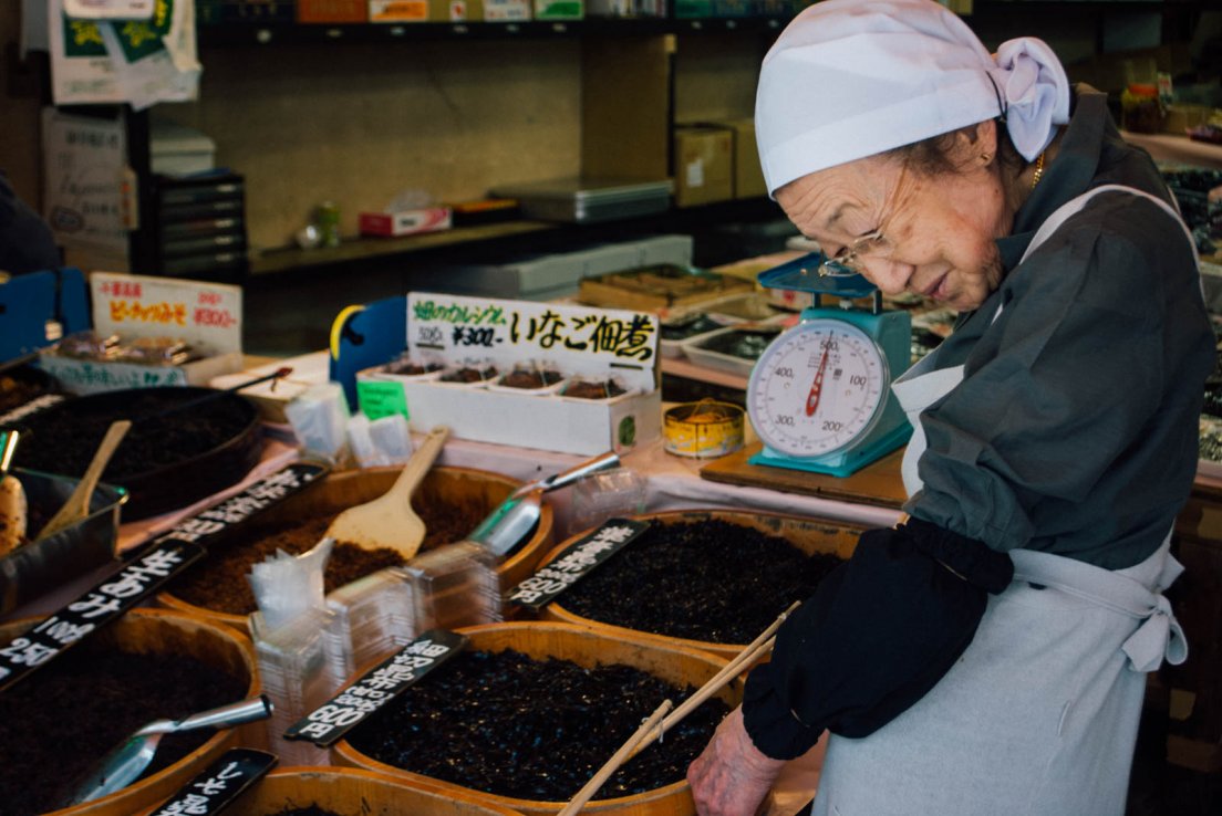 An elderly japanese lady standing by a stall displaying many varieties of seaweed