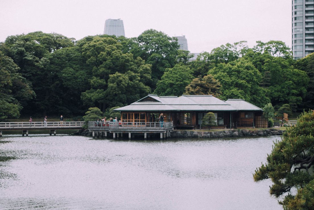 A tea house in the middle of a pond, bordered by tall skyscrapers