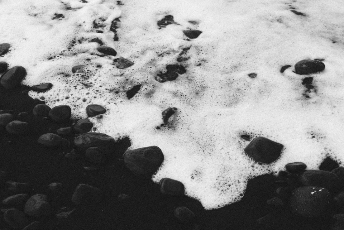 Black and white photograph of foam by a black sand sea shore